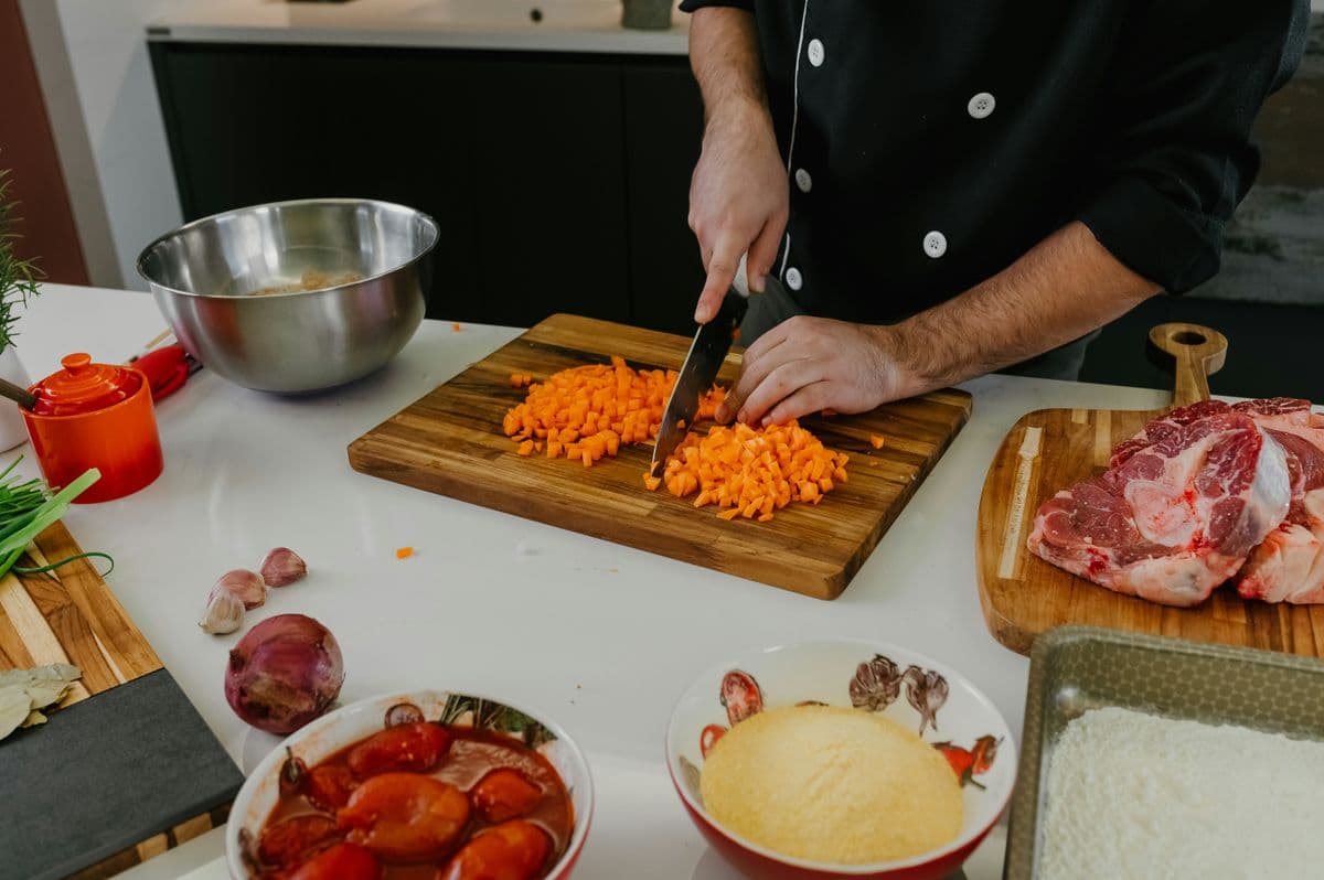 Dicing carrots on a wooden chopping board surrounded by meat and vegetables for a batch cooking session