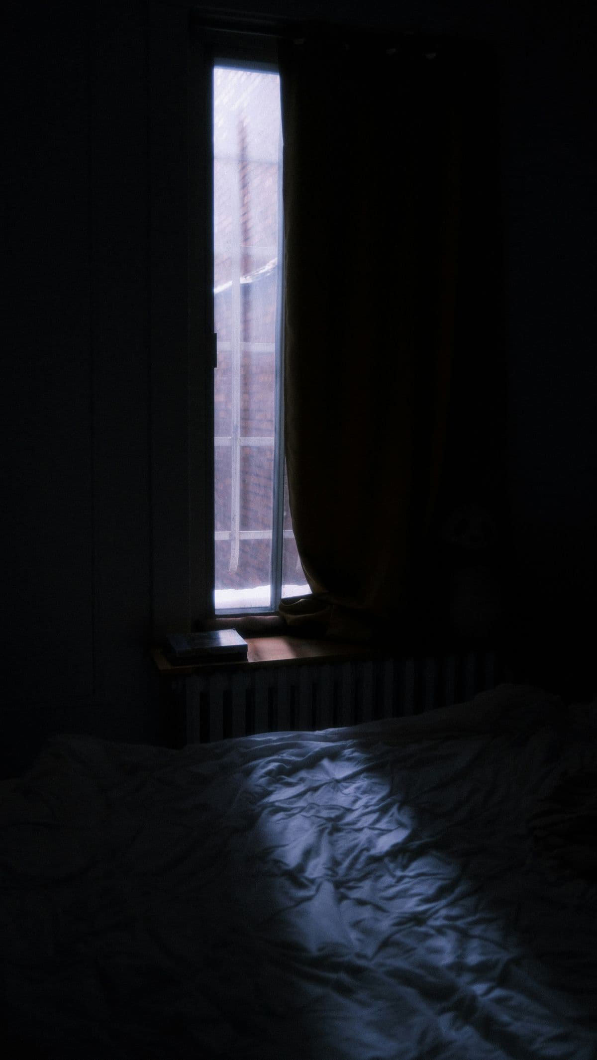 Dark bedroom with light filtering through a window onto rumpled bedsheets — the reality of daytime sleep for night shift workers